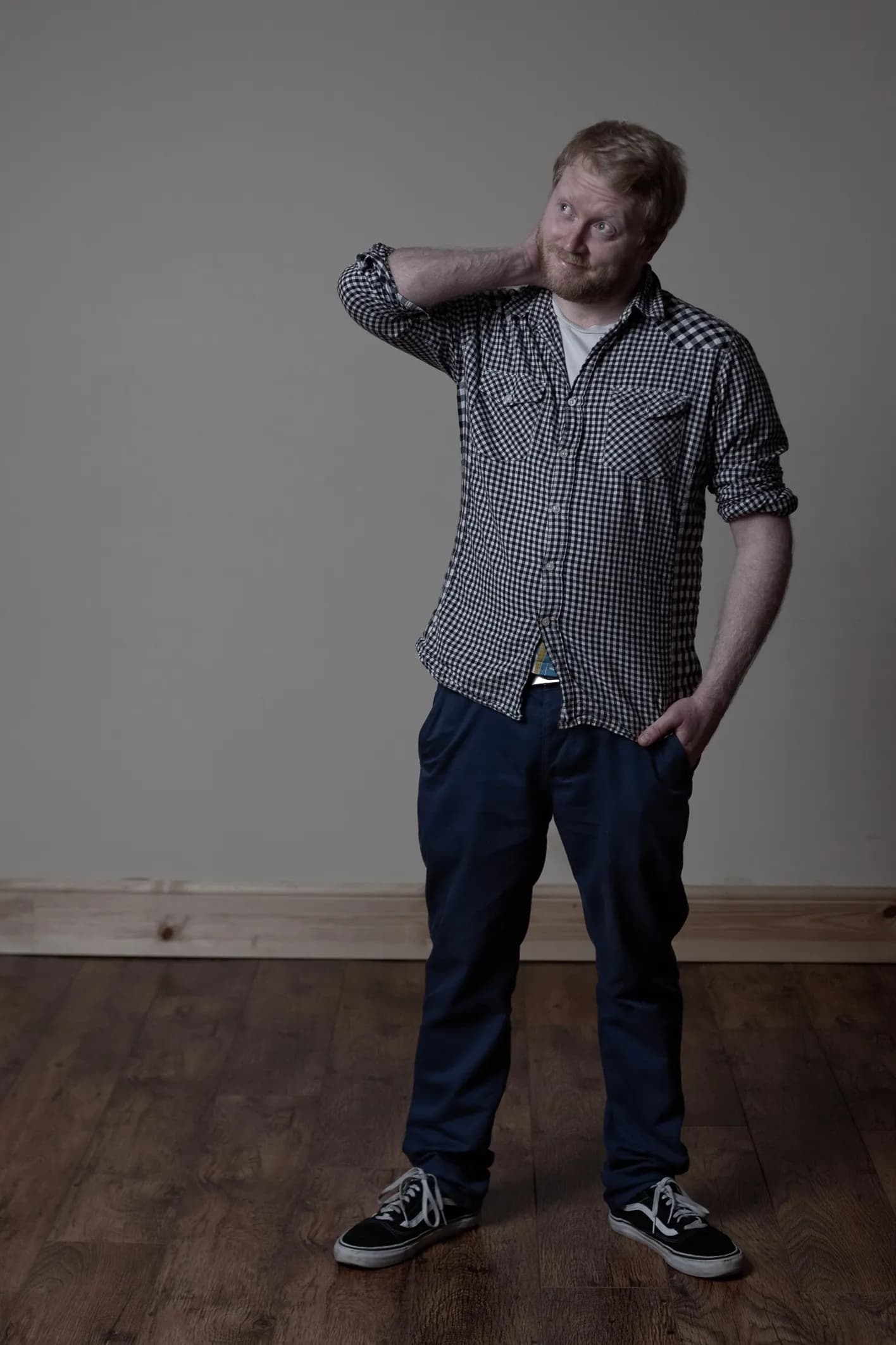 A man with a ginger beard, wearing a plaid shirt and dark blue trousers, stands with one hand scratching his neck, looking up and to the right.
