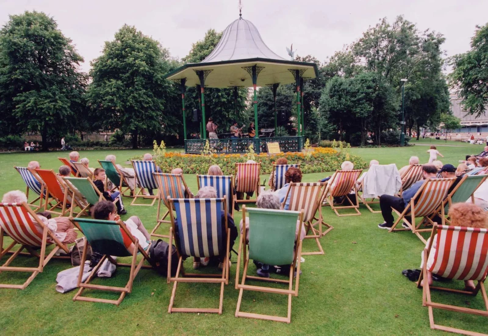 A group of people sitting in colorful striped deck chairs on a green lawn, facing a bandstand where musicians are performing.