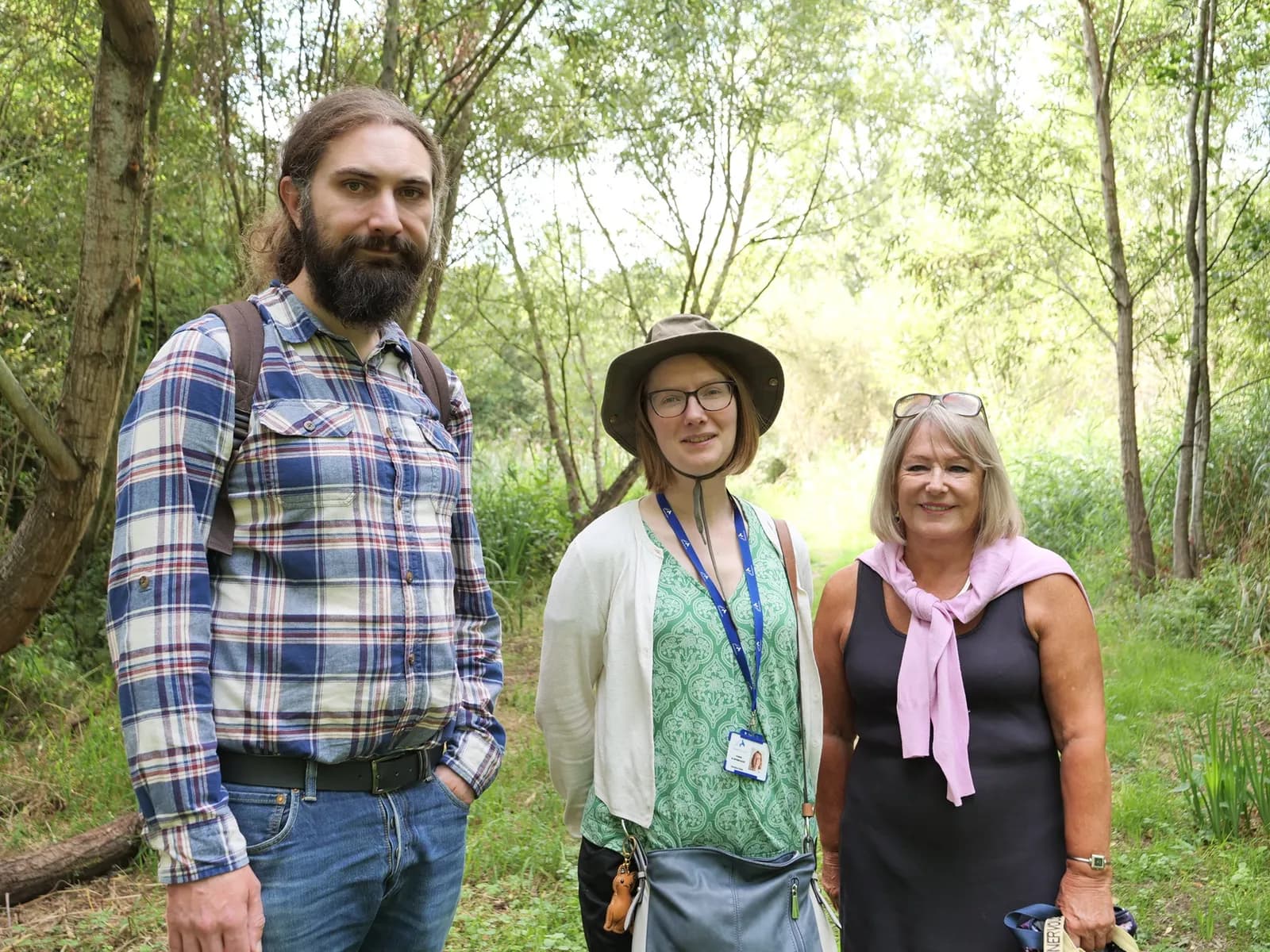 Three people, two women and one man, stand together on a dirt path in a wooded area.