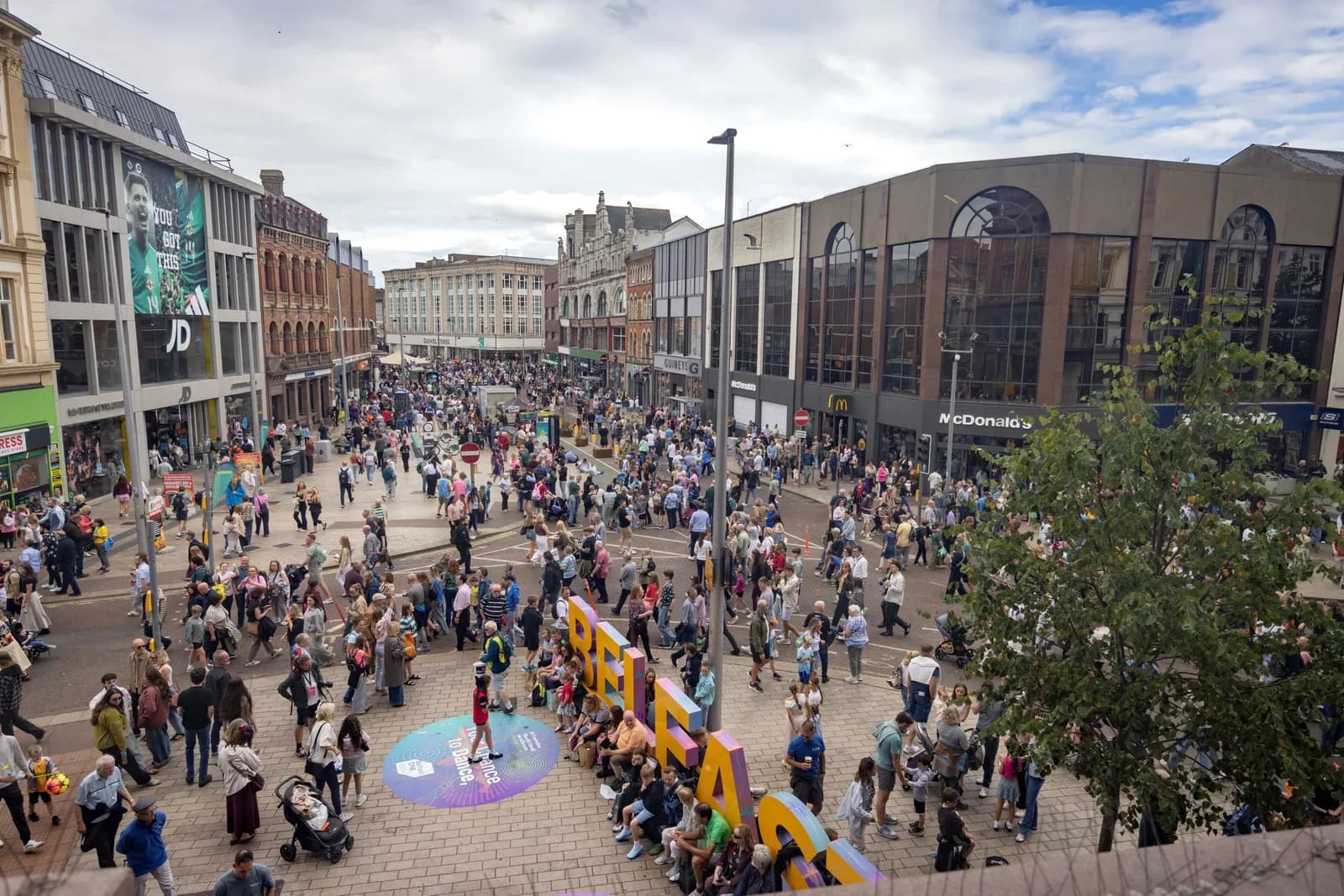 A wide shot from above shows a bustling city street filled with many people walking between shops, with large colorful letters spelling "BELFAST" visible in the foreground.