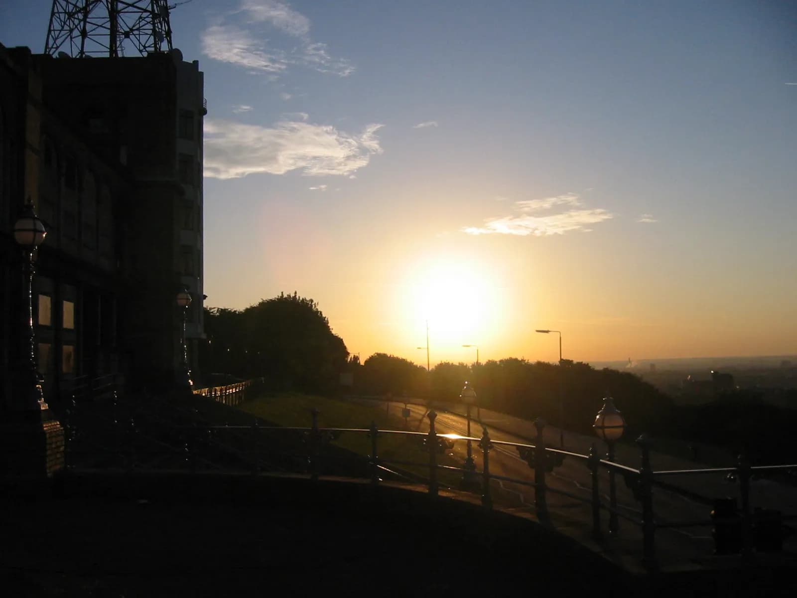 A beautiful sunset over hills and a distant city, with a large, dark building and decorative railings in the foreground.