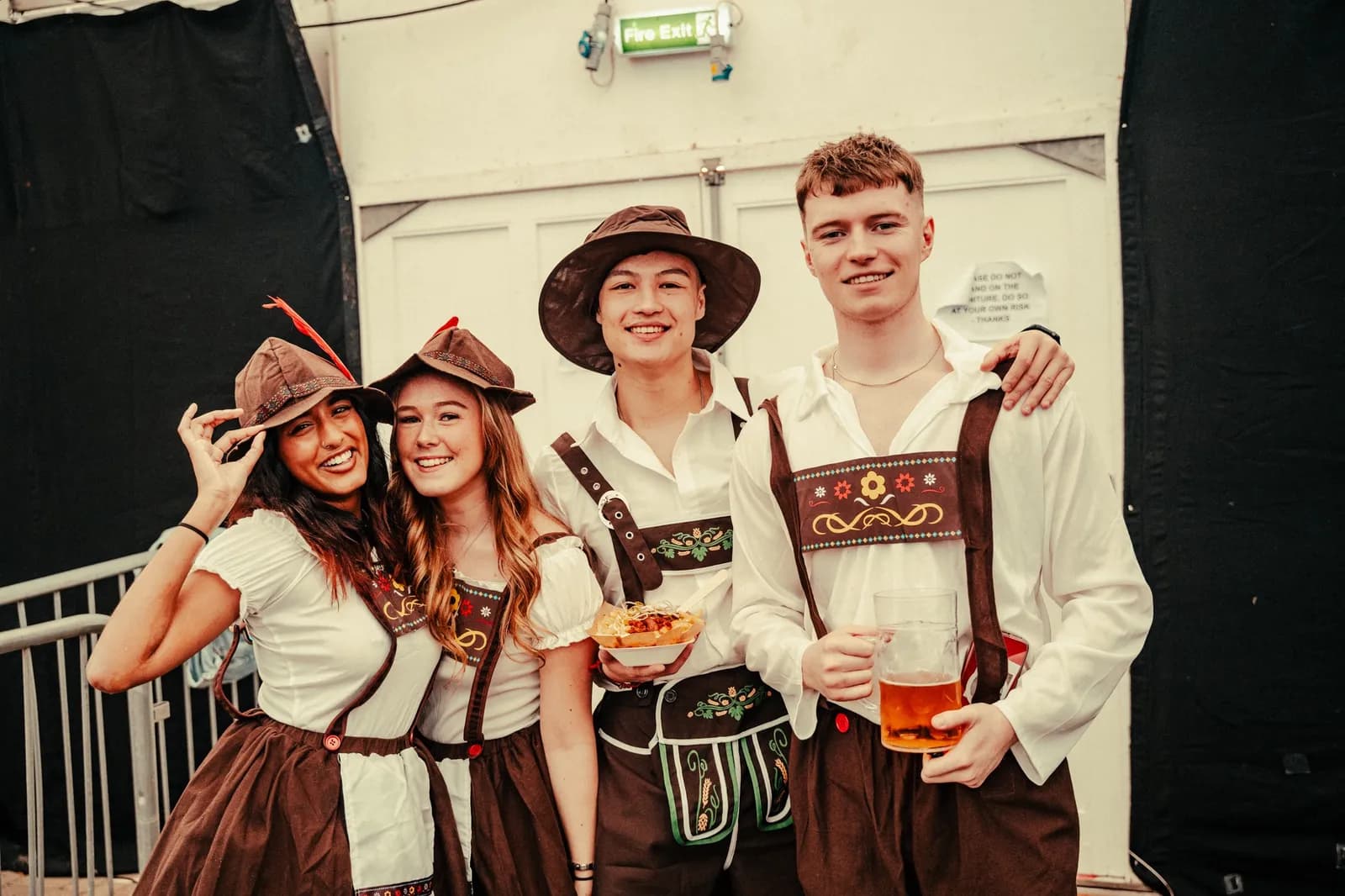 Four young adults, two men and two women, dressed in traditional Oktoberfest attire, smiling at the camera, with the man on the right holding a beer stein and the man in the middle holding a plate of food.