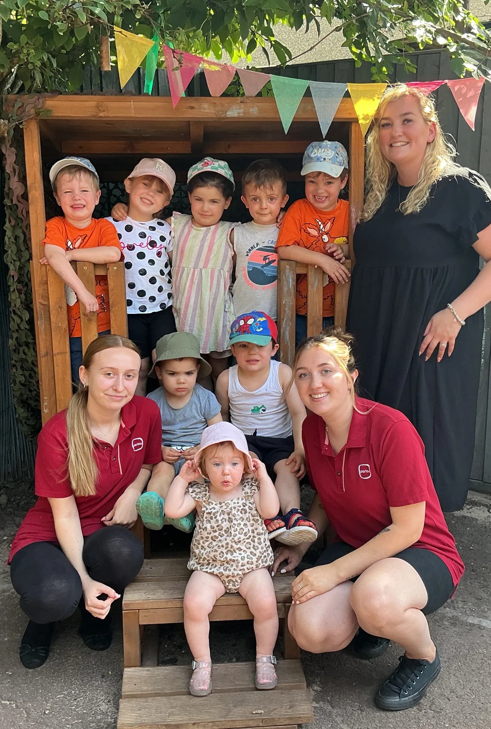A group of children and adults pose together in front of a wooden play structure decorated with colorful bunting.