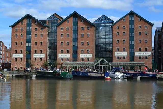 Several red brick buildings with dark windows and glass facades stand along a murky river where multiple long, narrow boats are docked, with people visible on some boats and along the riverside.
