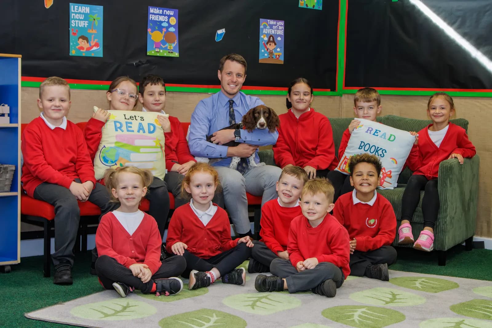 A man in a blue shirt sits on a red sofa with a brown dog in his arms, surrounded by ten children in red sweaters, all looking at the camera and smiling, some holding pillows that say "Let's READ" and "READ BOOKS".