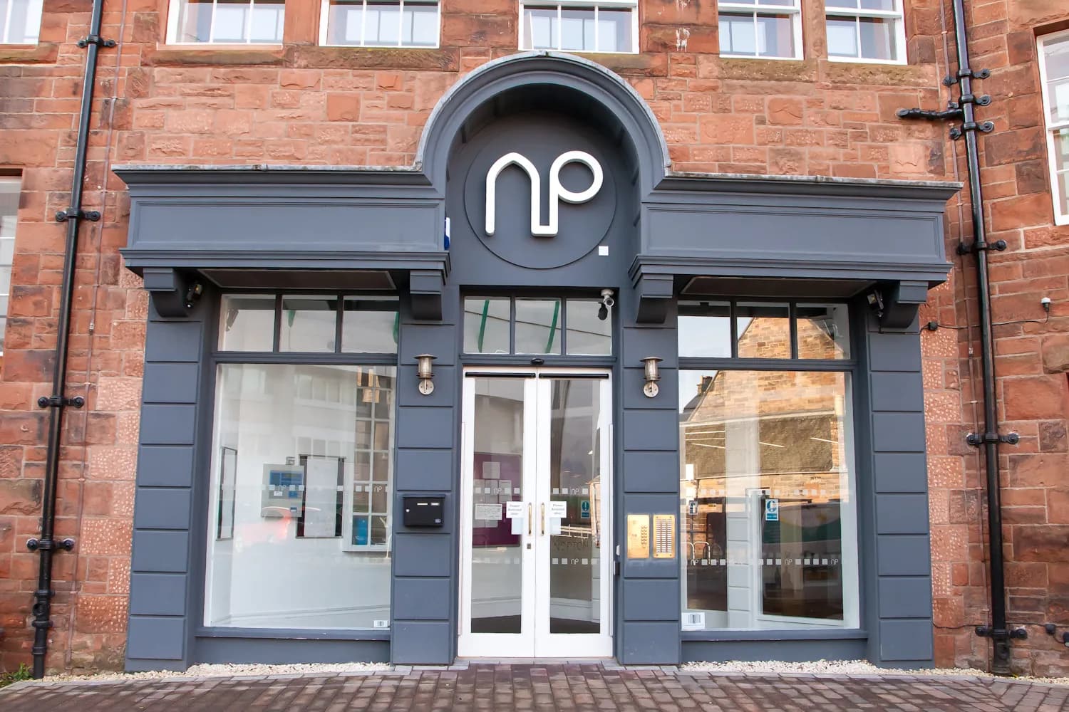 A storefront with a dark gray facade, large glass windows, and a white double door, set against a red brick building with an arched sign above the entrance.