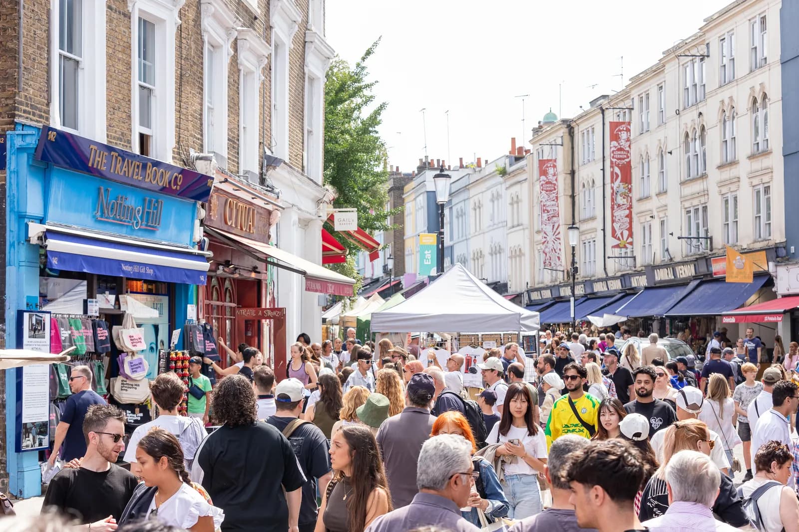 A busy street market scene with throngs of peoplewalking between storefronts and vendor stalls under a bright sky.