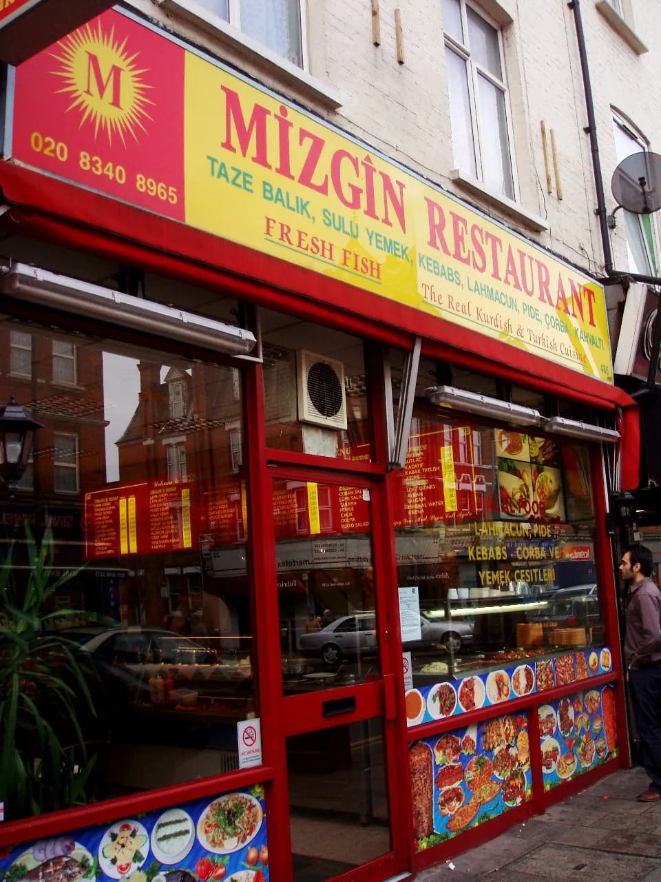 A man stands outside of Mizgin Restaurant, a Turkish and Kurdish establishment with a red and yellow sign and windows displaying food.
