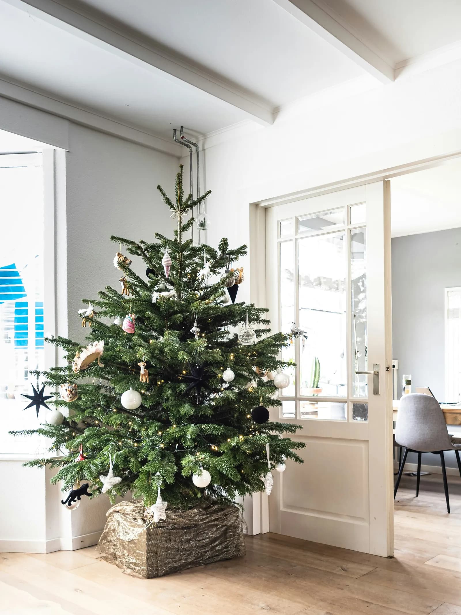 A decorated Christmas tree with various ornaments and lights stands in a room with a light wooden floor, next to a white door partially open to another room.