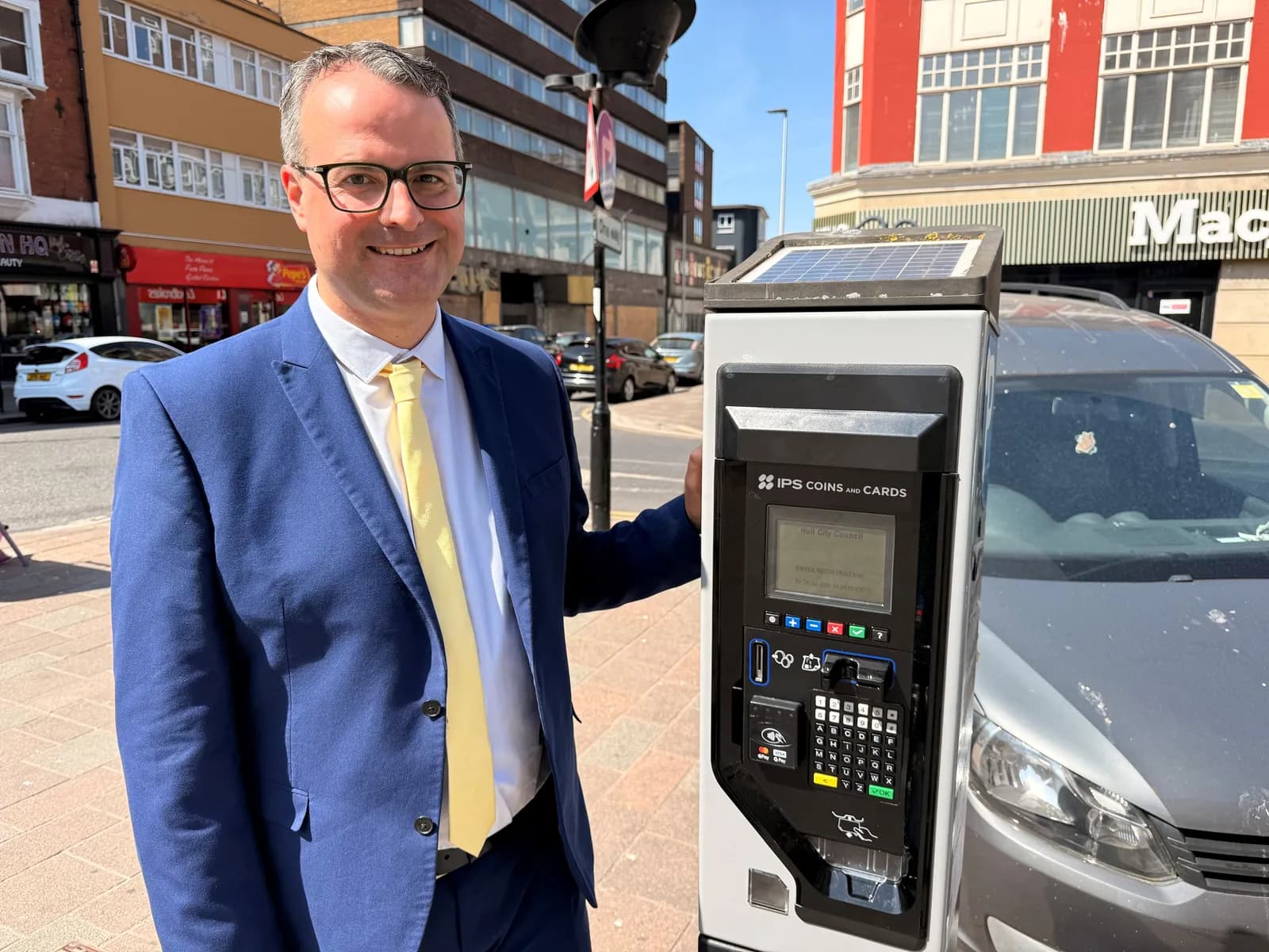 A man in a blue suit and yellow tie stands smiling next to a parking meter on a city street.