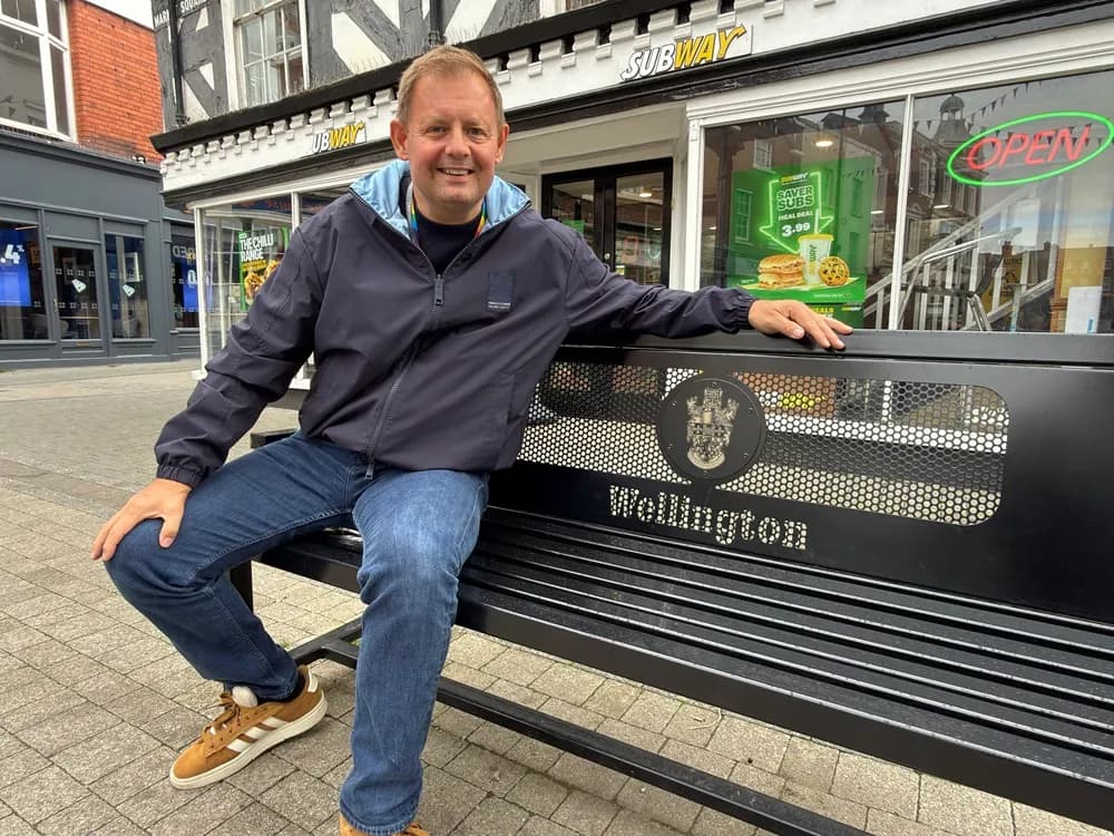 A man in a navy jacket and jeans sits on a black bench with "Wellington" inscribed on it, in front of a Subway restaurant.