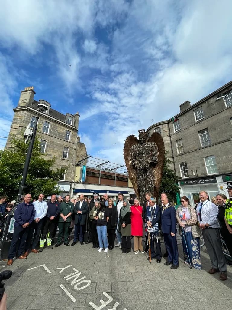 A group of diverse individuals, some in uniforms and suits, gather in front of a large, winged, metallic sculpture under a cloudy sky.
