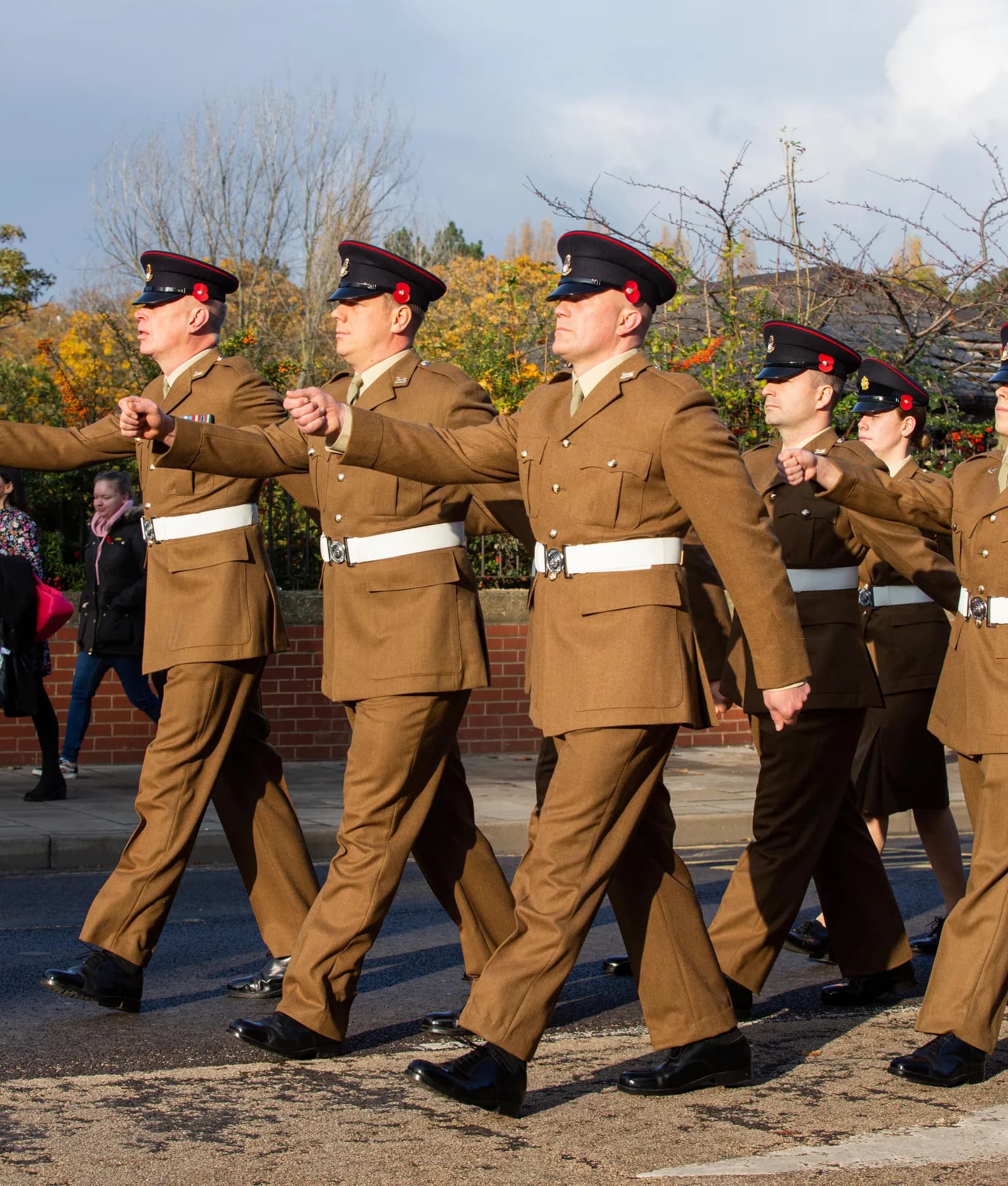 Several soldiers in brown uniforms with white belts and poppy pins march in formation down a street, with onlookers in the background.
