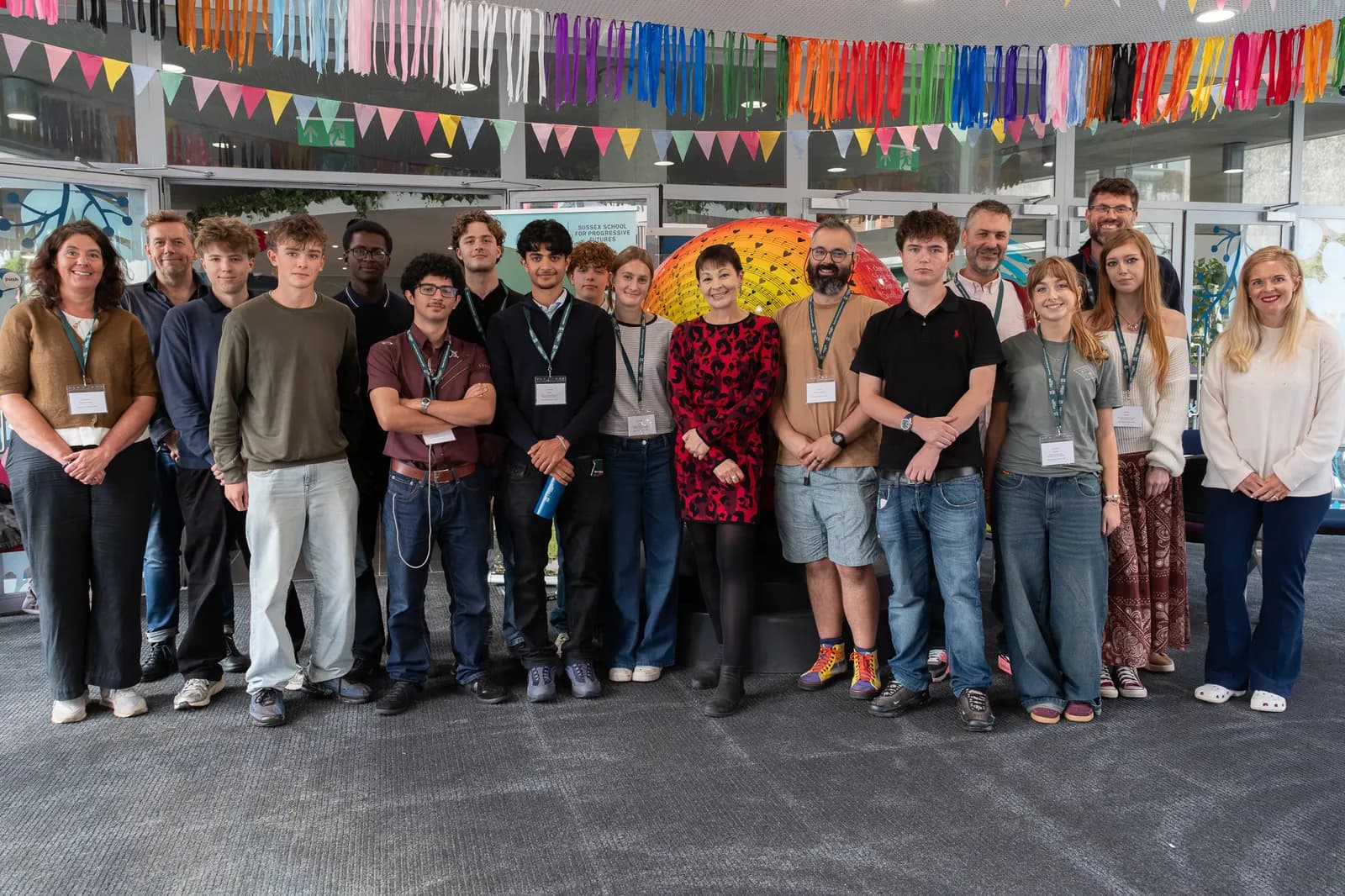 A group of diverse people, some with lanyards, standing indoors under colorful decorations, with an umbrella-like art piece behind them.