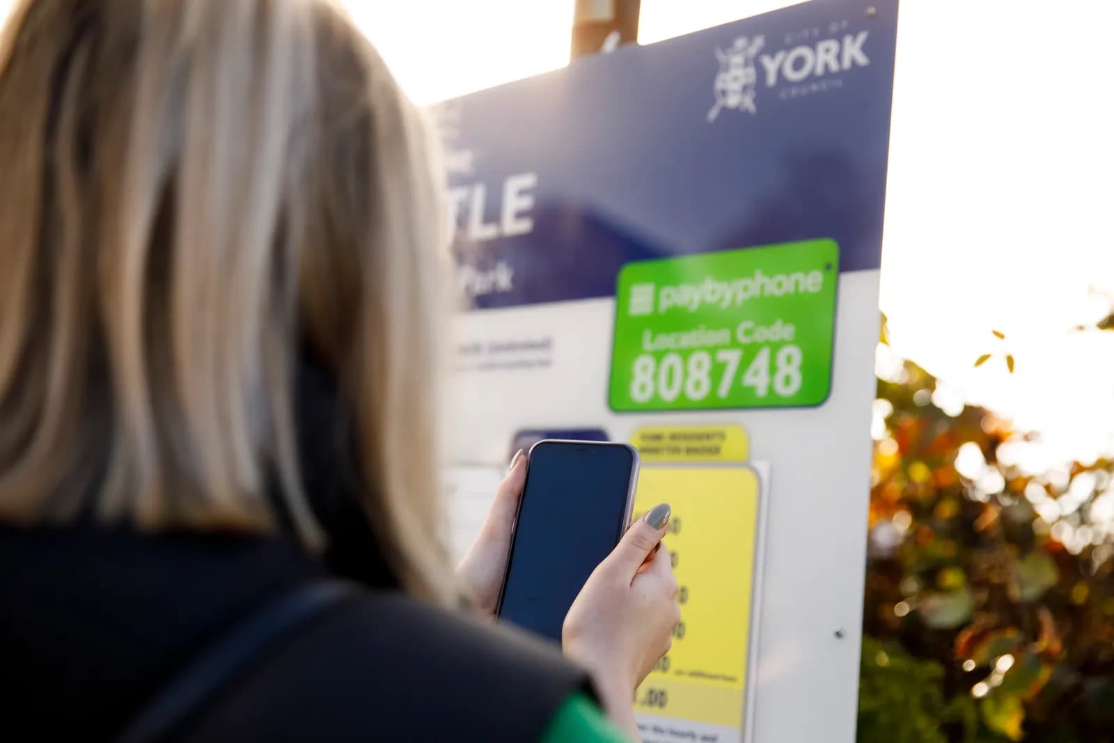 A person with blonde hair seen from behind holds a smartphone to pay using a pay-by-phone service at a parking meter with location code 808748, with sunlight and autumn foliage in the background.