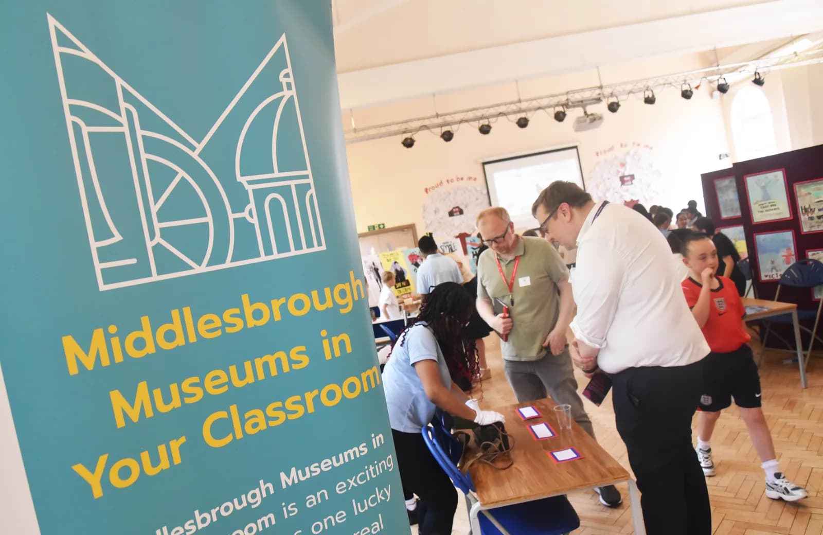Several people are gathered in a room, with a large teal banner promoting 'Middlesbrough Museums in Your Classroom' prominently displayed on the left.