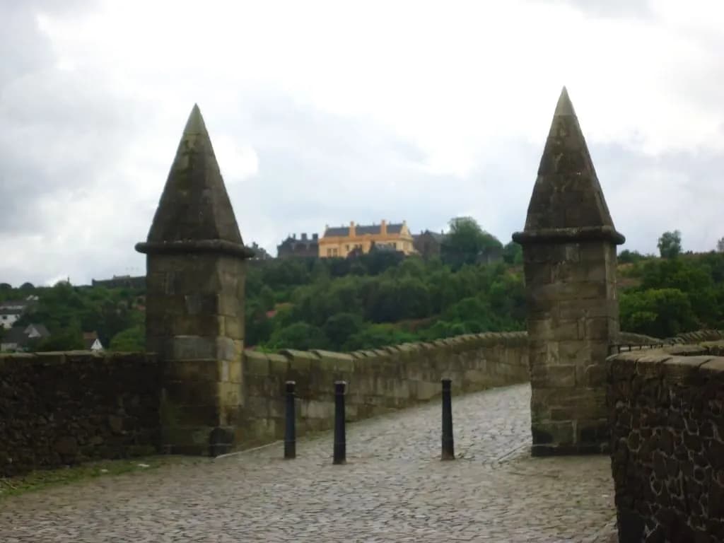 A view through two stone obelisks flanking either side of a cobbled path, revealing a yellow castle perched on a lush green hillside under a cloudy sky.