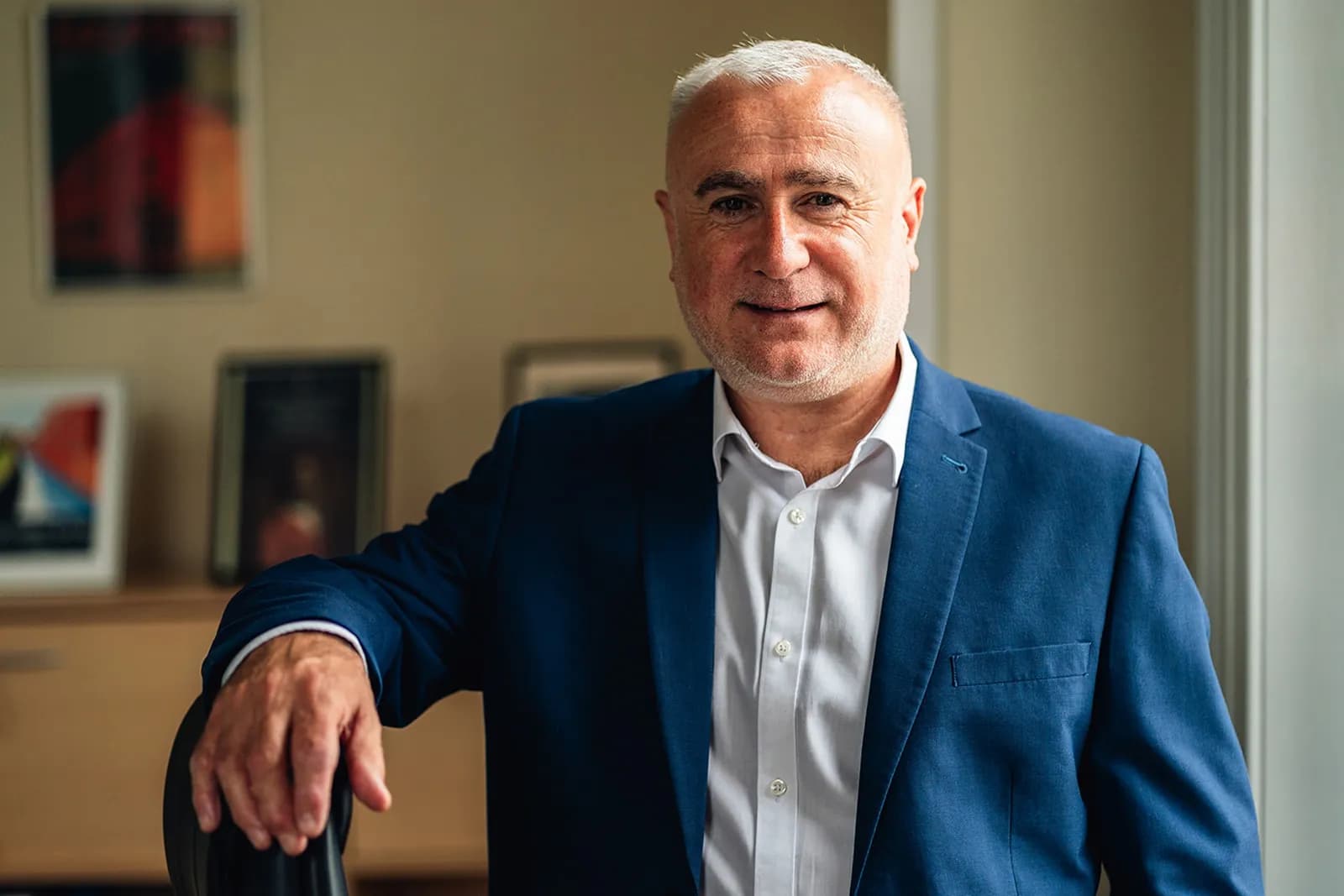 A man with a short white beard and white hair, wearing a white shirt and a blue blazer, leans on a chair arm and smiles slightly at the viewer.