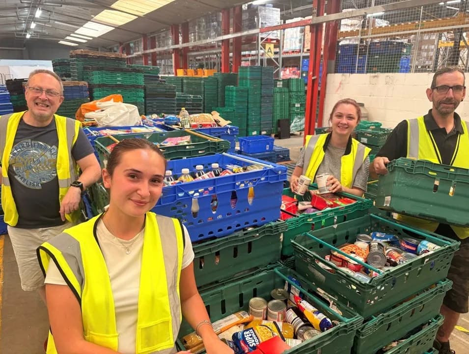 Four volunteers in high-visibility vests smile while sorting food items into crates within a warehouse setting.