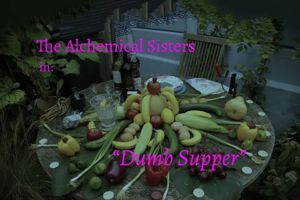 A wooden table is covered with an arrangement of fruits, vegetables, and eggs, centered around a fruit creation resembling a person, with text overlayed that reads "The Alchemical Sisters in: 'Dumb Supper'".