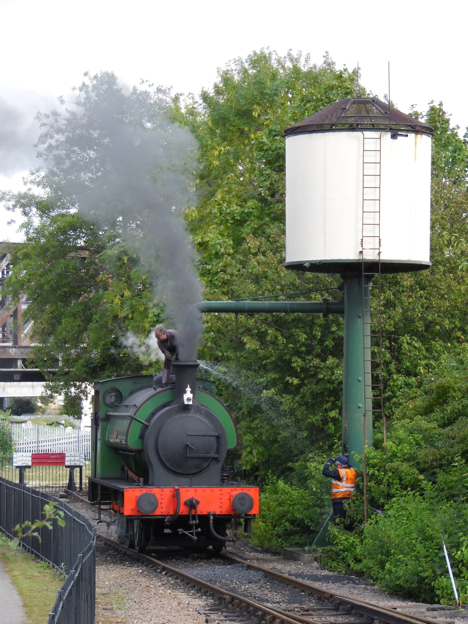 A man is working on top of a green and black steam locomotive that is emitting a large plume of smoke, while another man in an orange vest stands next to a tall white water tower in a leafy, outdoor setting.