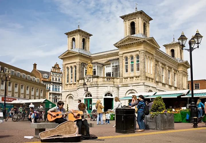 Two young men play acoustic guitars next to an open guitar case on a stone bench in a bustling town square with a grand stone building featuring towers and a golden statue in the background.