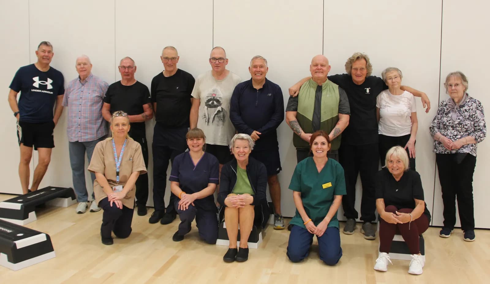 A group of older adults and healthcare professionals posing for a picture in a brightly lit room with wooden floors and white walls, with some exercise steps in the foreground.