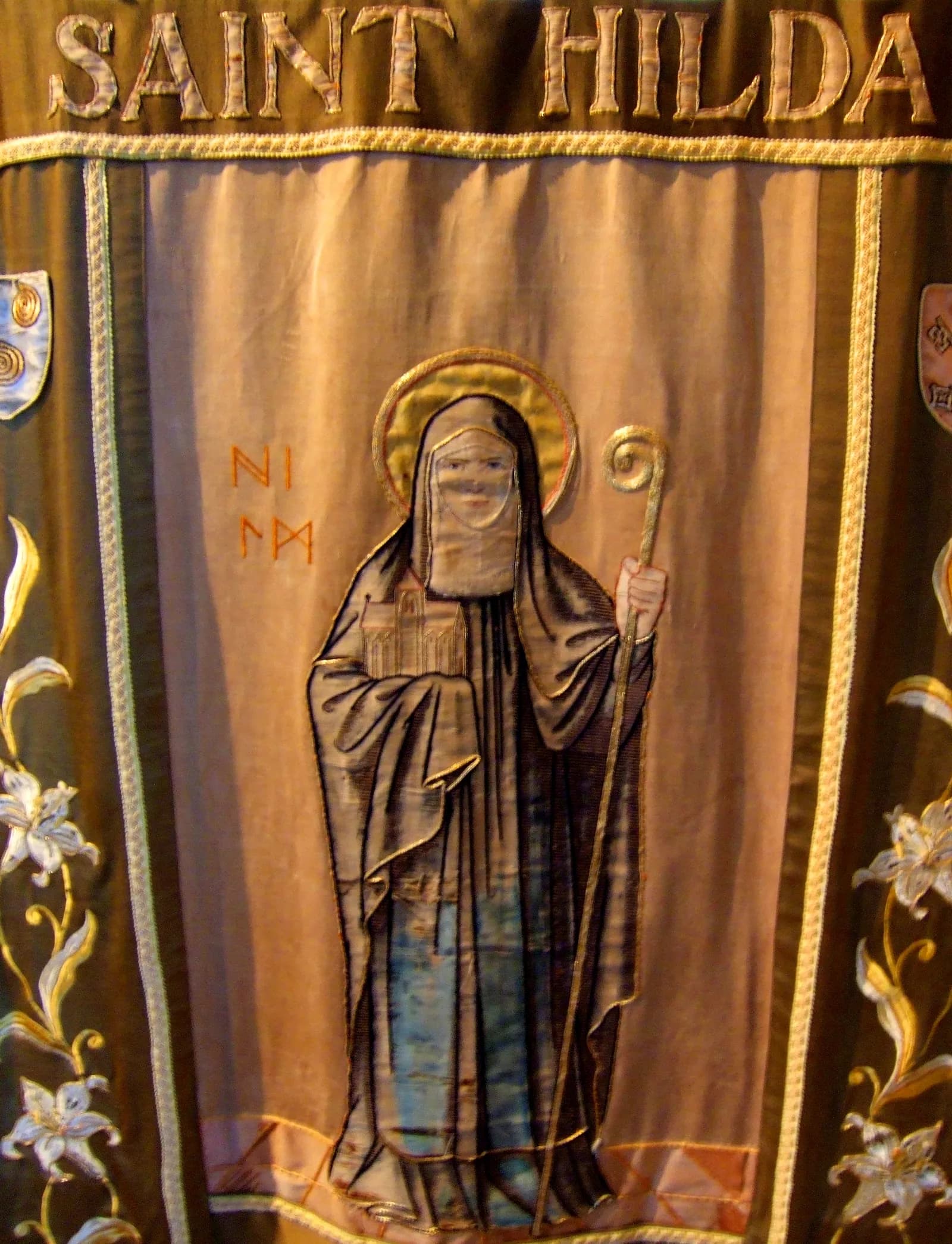 A tapestry banner features an embroidered depiction of Saint Hilda with a halo, holding a book and a staff, and the words "SAINT HILDA" stitched above.