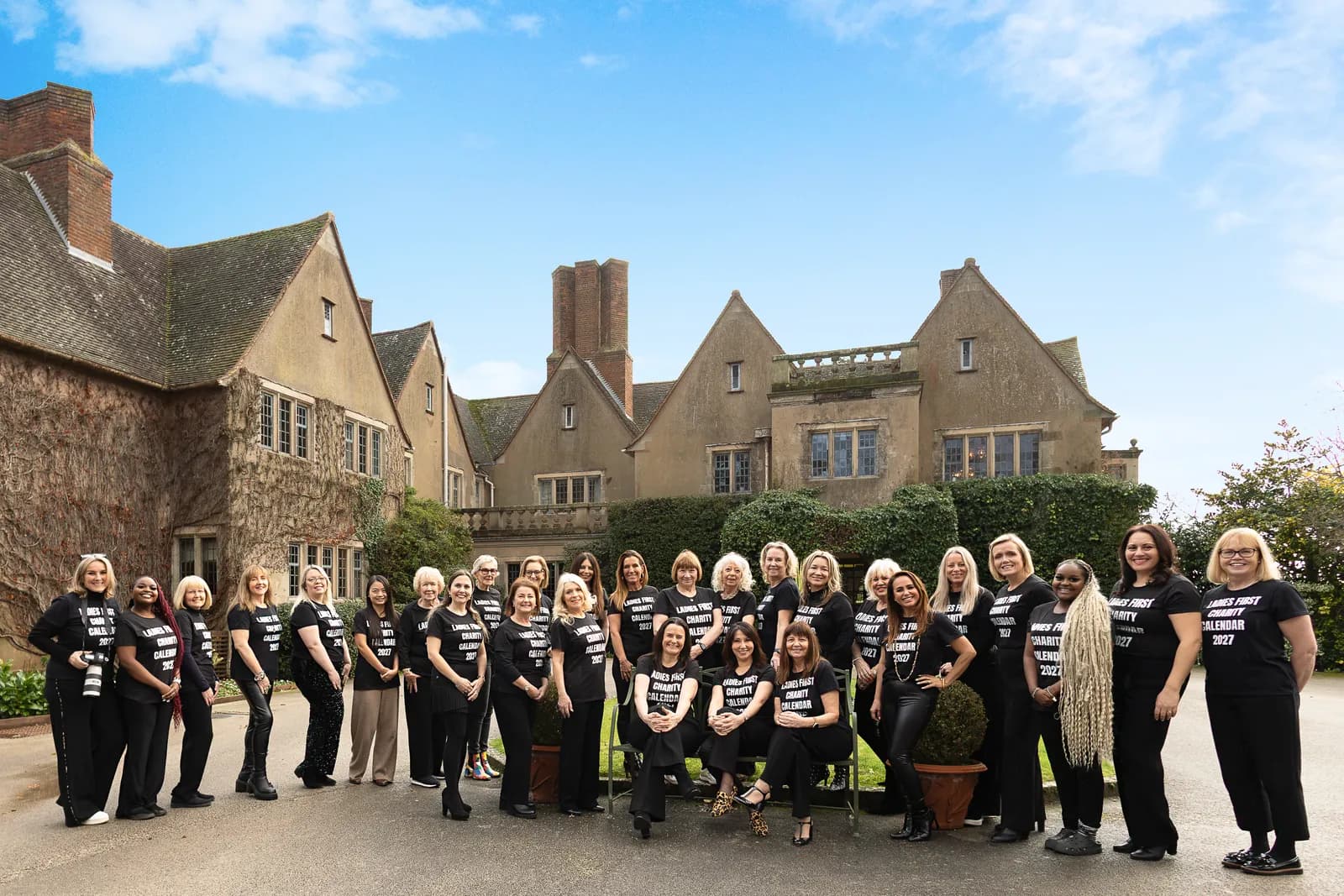 A group of women in black t-shirts and pants stand and sit outside a large, ivy-covered stone building with a clear blue sky above.