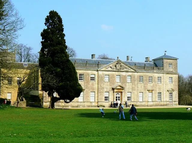 A grand, multi-story stone manor with a large green lawn in front, where people are playing and relaxing under a bright blue sky.