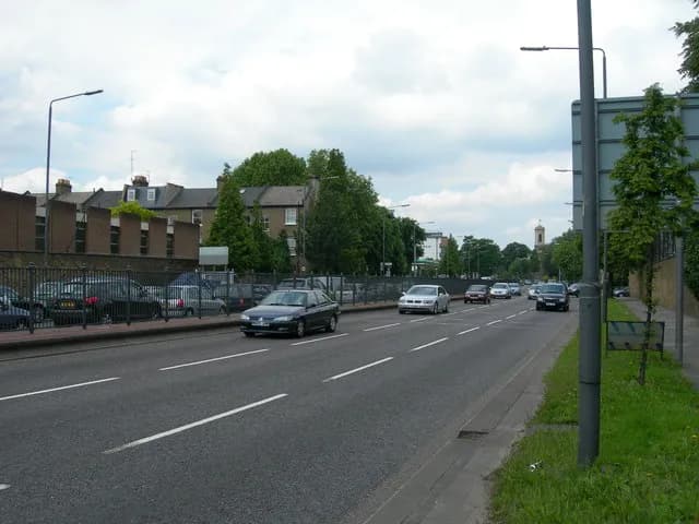 Cars drive along a multilane road with a black car in the foreground, alongside buildings and trees under a cloudy sky.