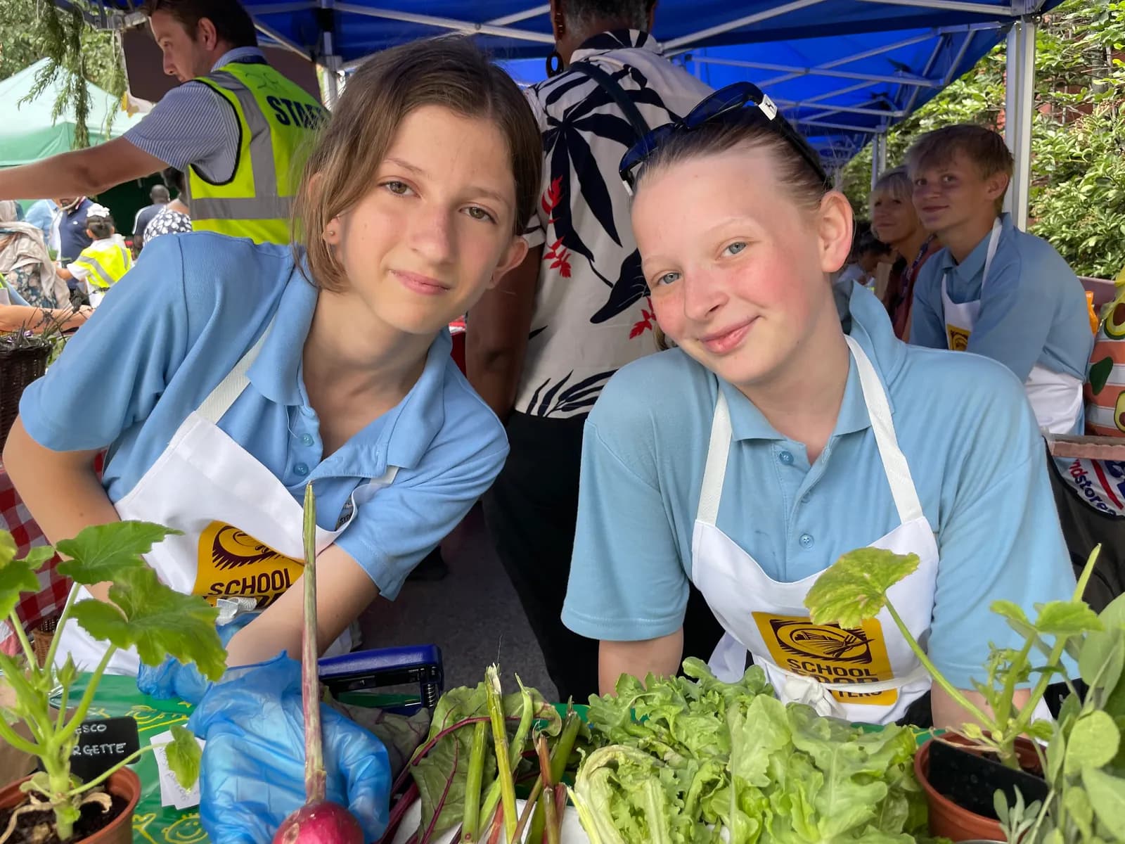 Two young girls in blue shirts and white aprons smile behind a stall filled with fresh produce and plants, with other people and market stalls in the background.