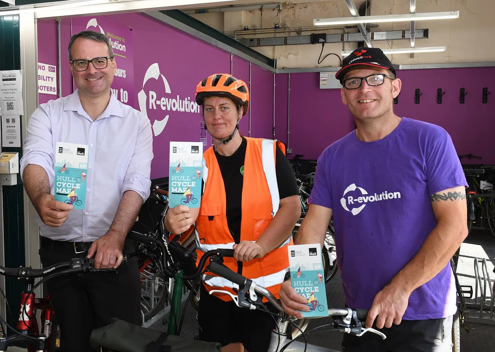 Three people, two men and one woman, stand behind bicycles, holding up Hull Cycle Maps, with a purple wall with "R-evolution" text in the background.
