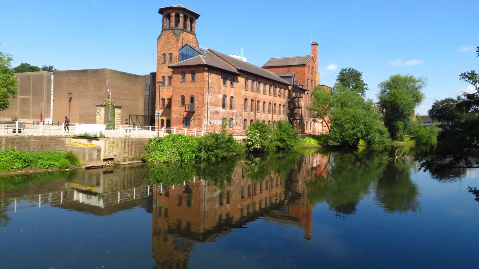 A historic brick factory building with a large tower stands by a river, with green trees and its reflection visible in the water.