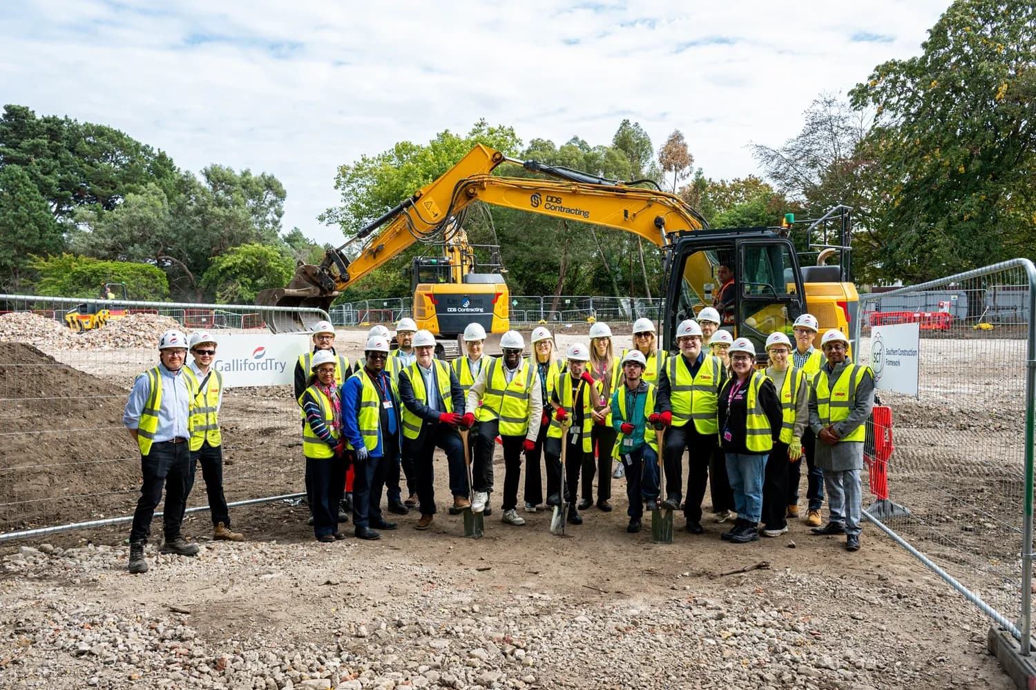 A diverse group of construction workers in hard hats and high-visibility vests stands in front of a yellow excavator at a construction site.