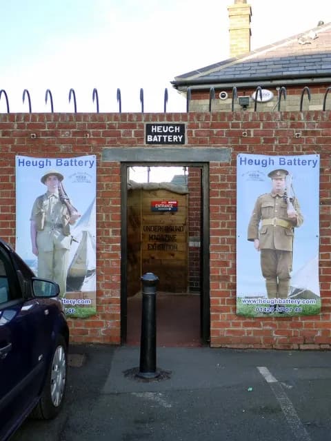 A brick building with an entrance to the 'Heuch Battery' museum, flanked by two banners featuring soldiers in historical uniforms, with a blue car in the foreground.