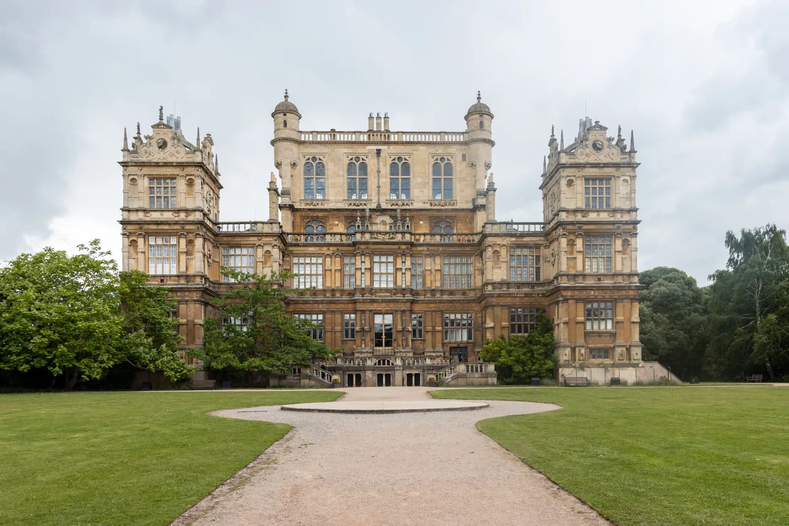 A grand, symmetrical, multi-story stately home with ornate architecture, framed by a green lawn and trees under a cloudy sky.