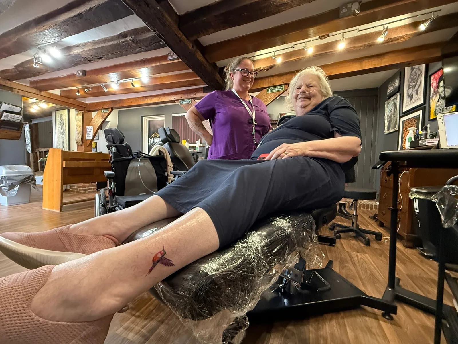 An elderly woman with a hummingbird tattoo on her leg smiles while seated in a tattoo parlor, with a tattoo artist standing behind her.