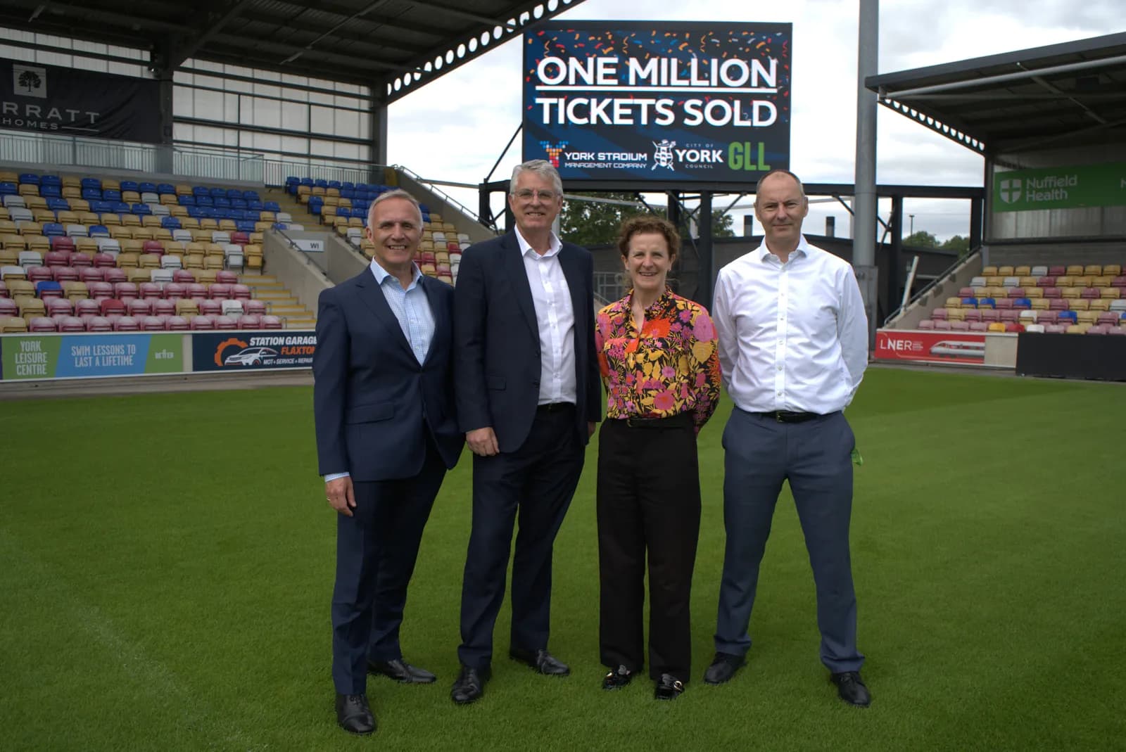 Four people, two men in suits, one woman in a floral top, and one man in a white shirt and dark trousers, stand on a green field in front of a large scoreboard that reads "ONE MILLION TICKETS SOLD" within a stadium.