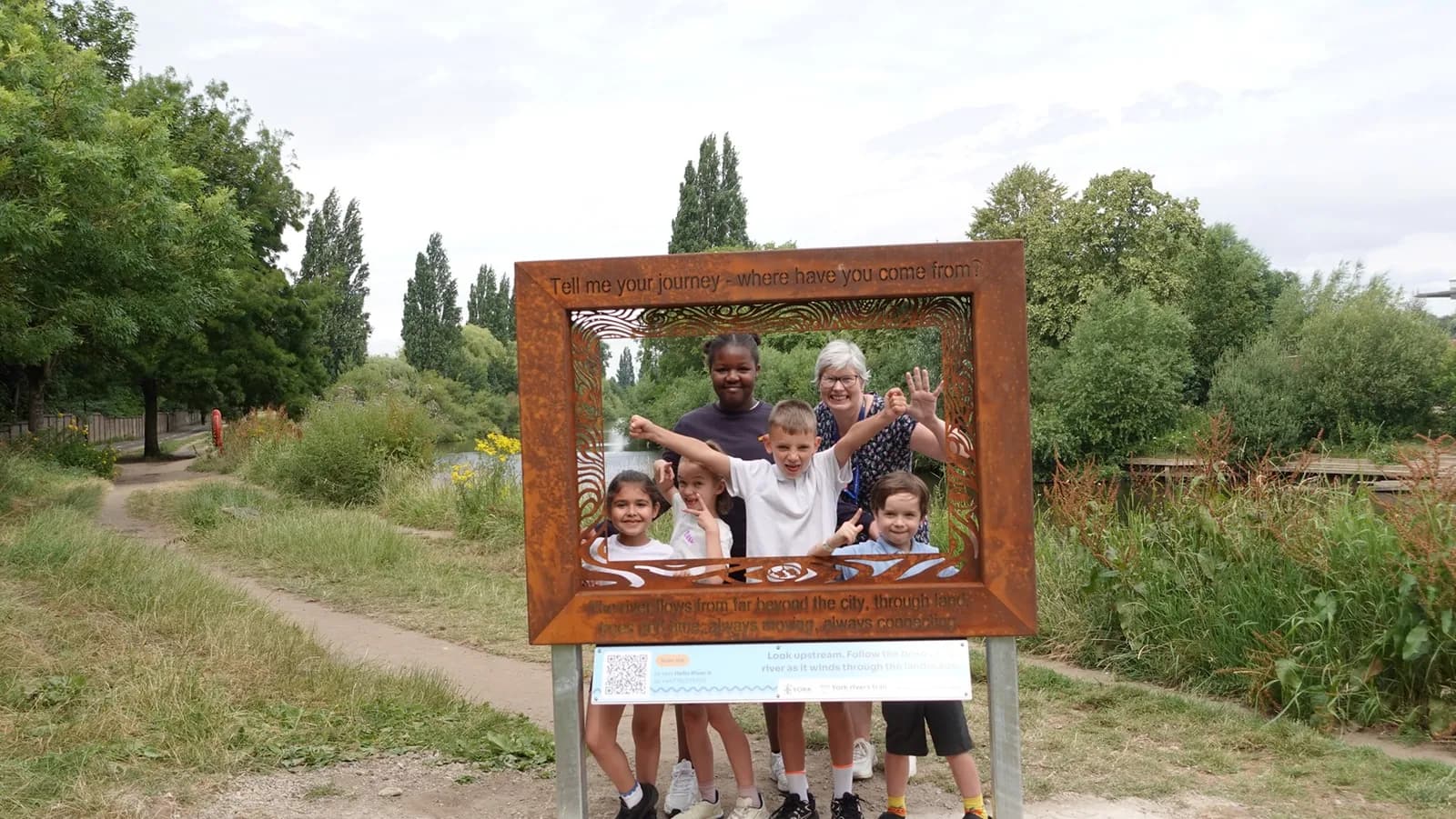 A group of six children and two adults pose behind a rustic metal frame with text overhead, smiling directly at the camera, with a view of a path and trees in the background.