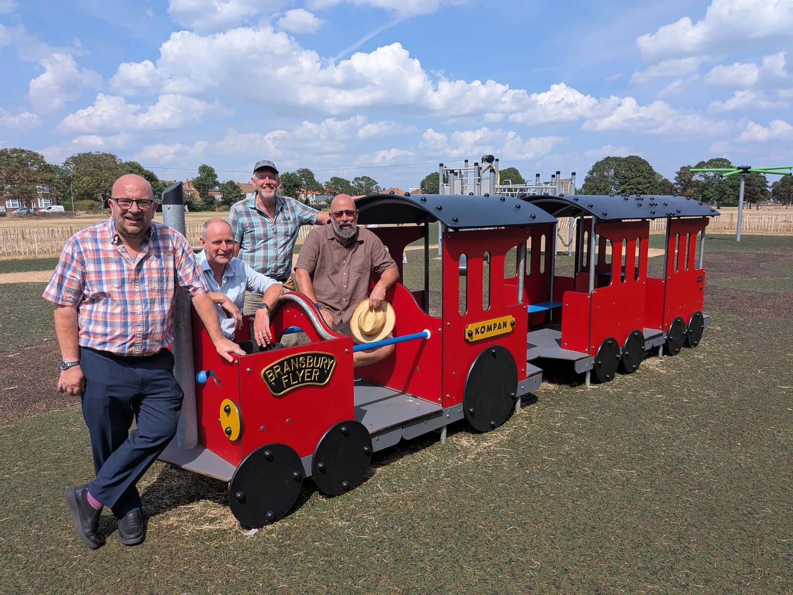 Four men posing with a red play train under a blue sky, with one man standing and three seated inside the train cars.