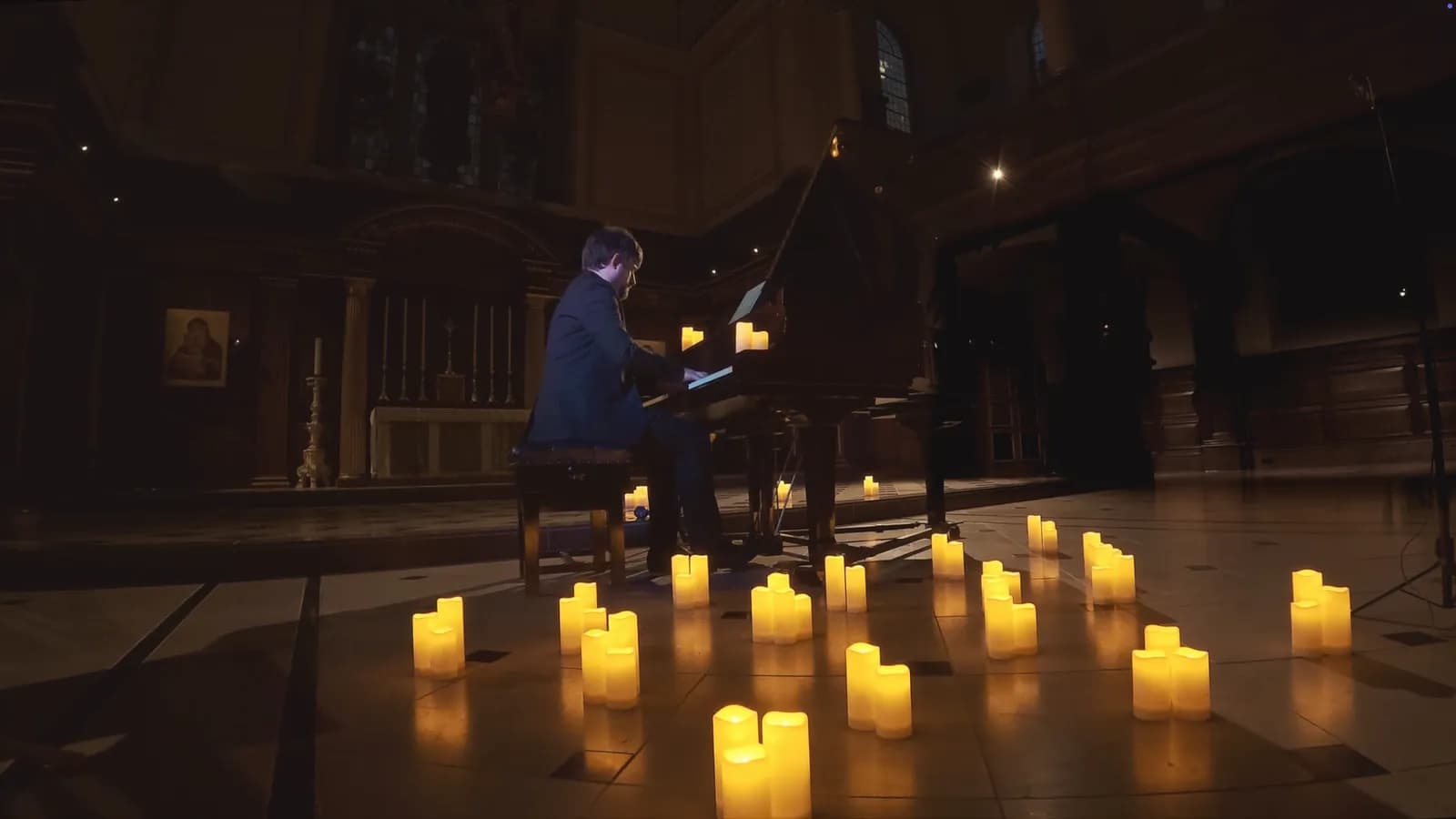 A man in a dark suit plays a grand piano in a dimly lit room, surrounded by numerous glowing pillar candles on the floor.