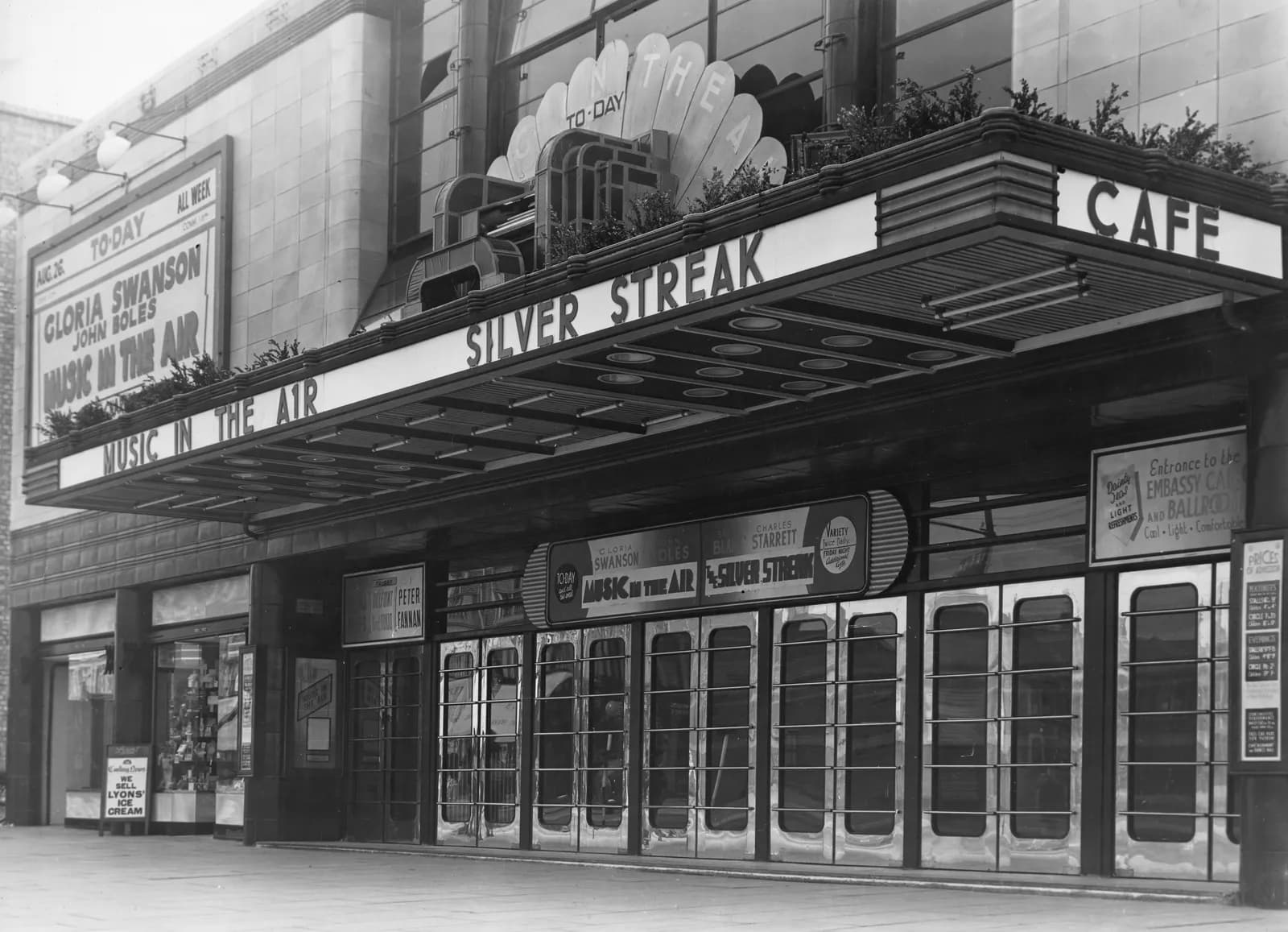 The black and white street view of a theatre entrance with an awning and signage showing the movie titles 'Silver Streak' and 'Music in the Air'.