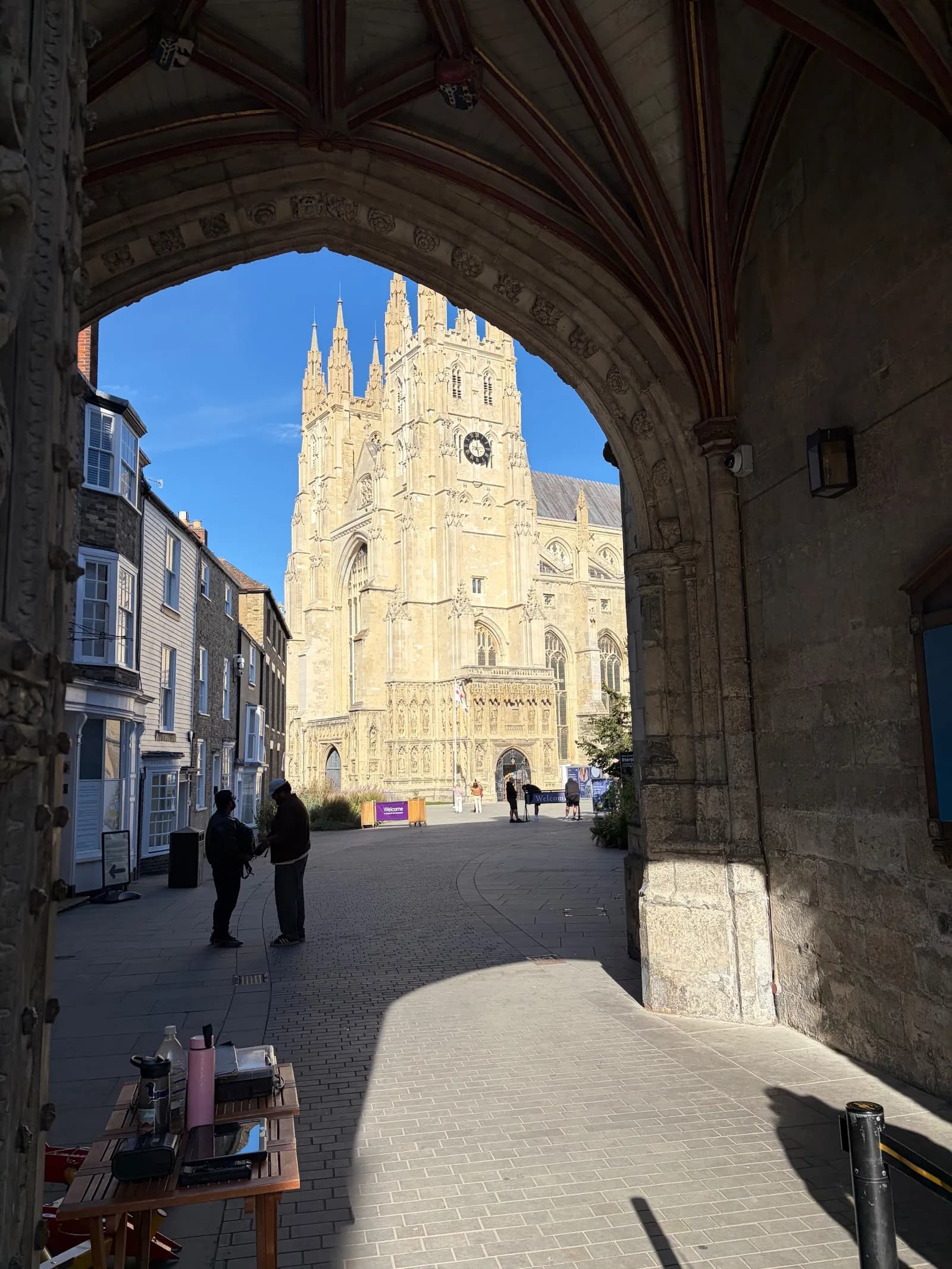 A grand cathedral with spires and a clock tower is visible through a dark, stone archway, with two people standing in the sunlit street below.