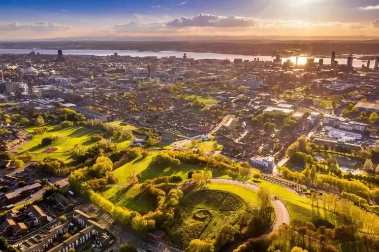 An aerial view of a sprawling city with green parks and a river in the background under a warm, sunny sky.