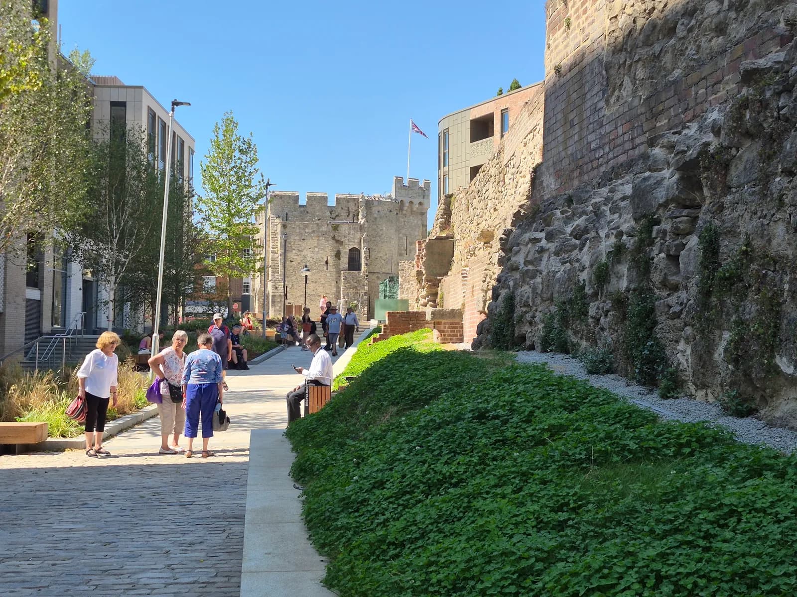 People walk and sit in a modern urban plaza with old stone castle walls on the right and contemporary buildings on the left.