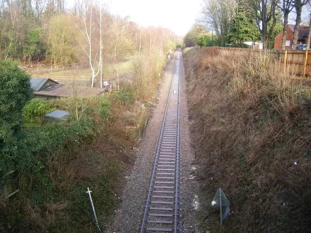 A long, straight railroad track extends into the distance, flanked on both sides by grassy embankments and trees, with a building visible on the left.