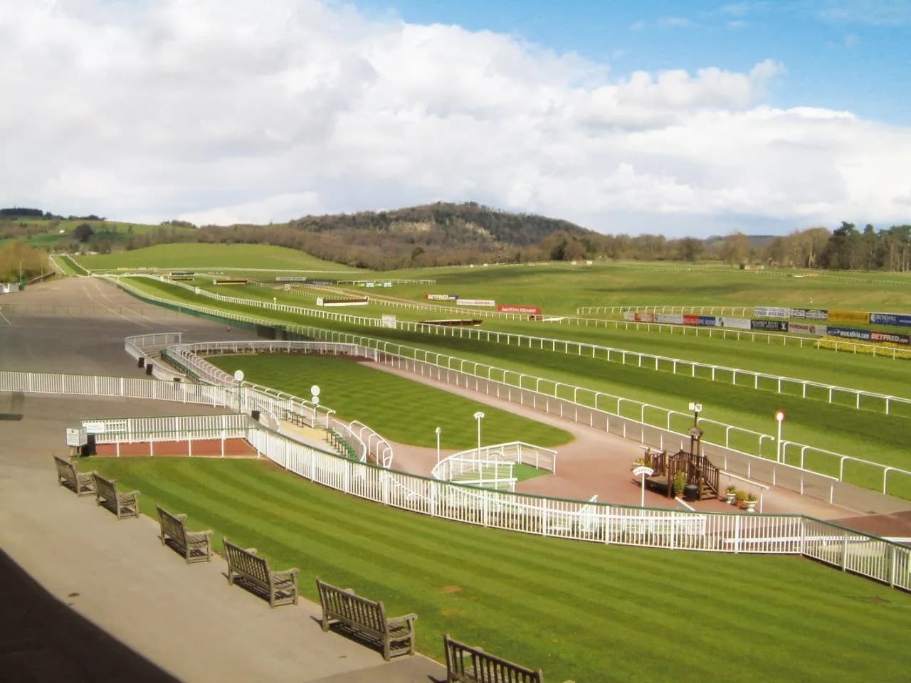 A wide shot of a green horse racing track and surrounding grassy fields under a blue sky with white clouds, with rolling hills in the background.