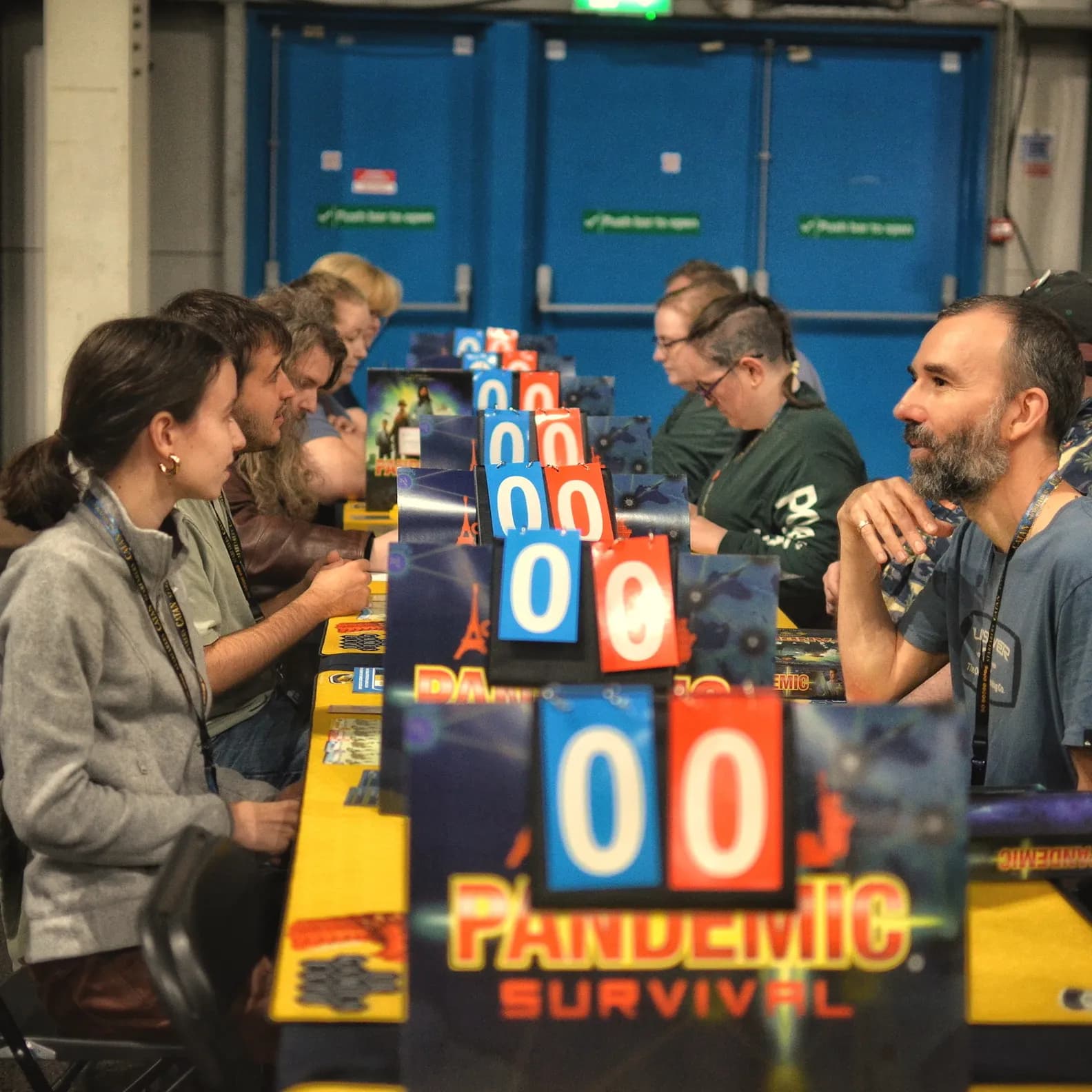 A group of people sitting at a long table covered with game boards and numbered display cards, focused intently on a board game in a brightly lit indoor setting.