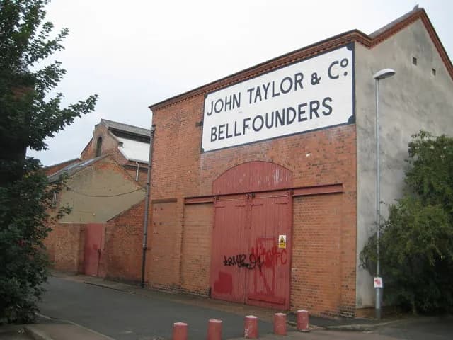 A historic brick building with a large white sign reading "JOHN TAYLOR & CO BELLFOUNDERS" above a prominent red gate, along a narrow street.